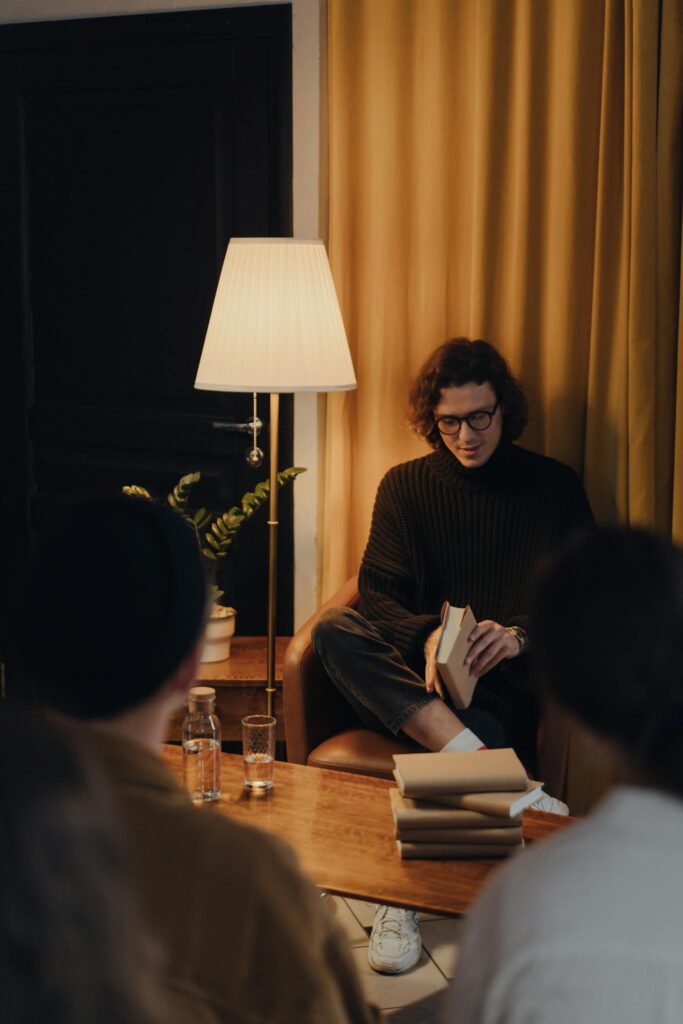 A man reads a book to a small group in a cozy, warmly lit bookstore setting.