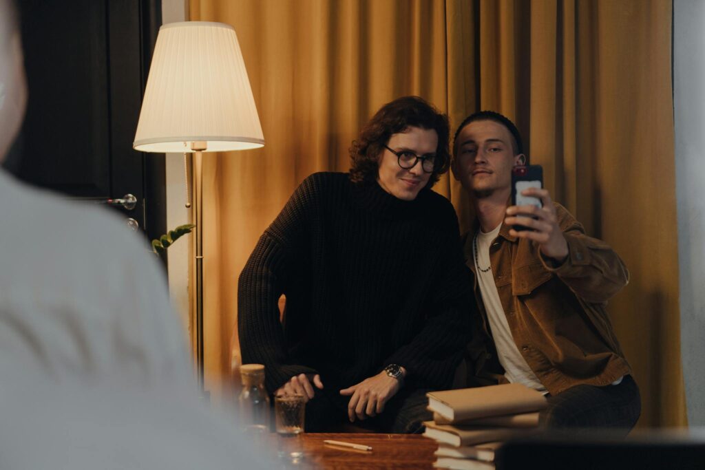 Two young men capture a selfie indoors with books stacked nearby, creating a cozy ambiance.